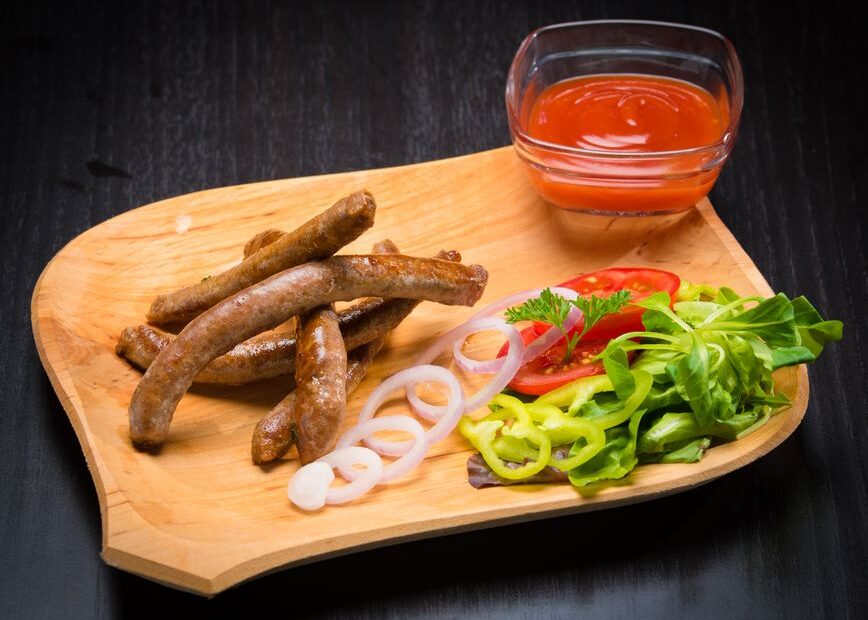 Grilled sausages with sliced vegetables and tomato sauce on a wooden plate.
